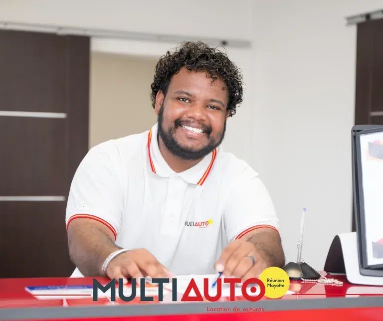 Agent Multi Auto souriant au comptoir d'une agence, prêt à accompagner un client qui souhaite préparer sa location de voiture à La Réunion